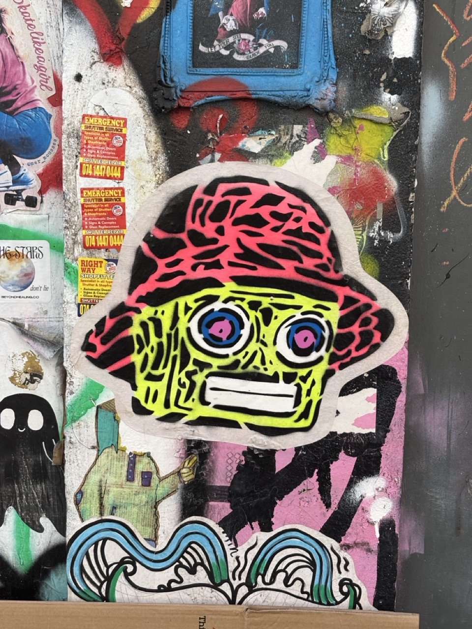 A photo of a street art paste-up. It appears to be a cartoon robot head wearing a bucket hat, in a colour scheme of neon yellow and neon reddish-pink.