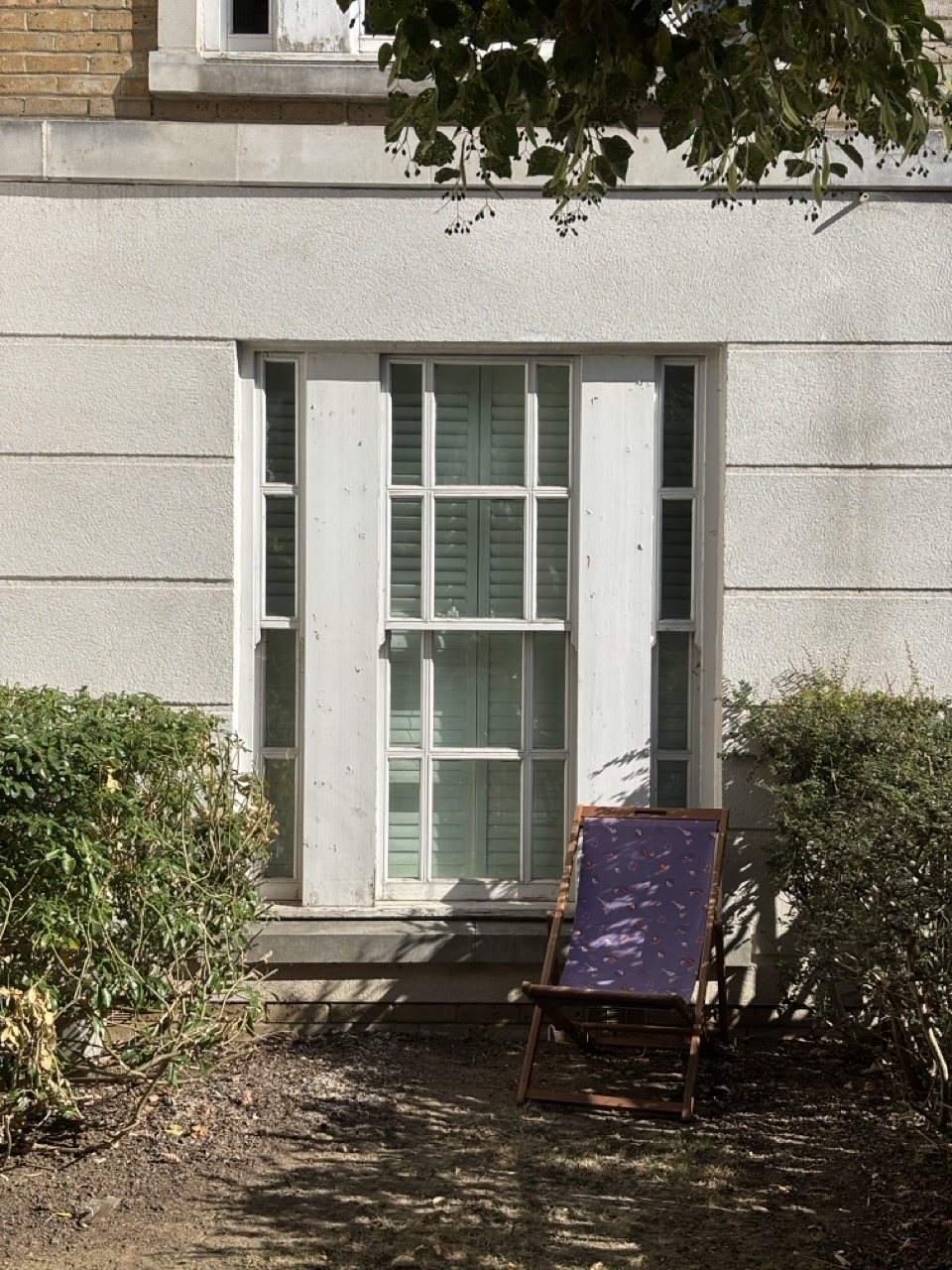 A photo of a seat made out of purple cloth and a wooden frame, outside the window of a residential building, in-between two plants and partially shaded