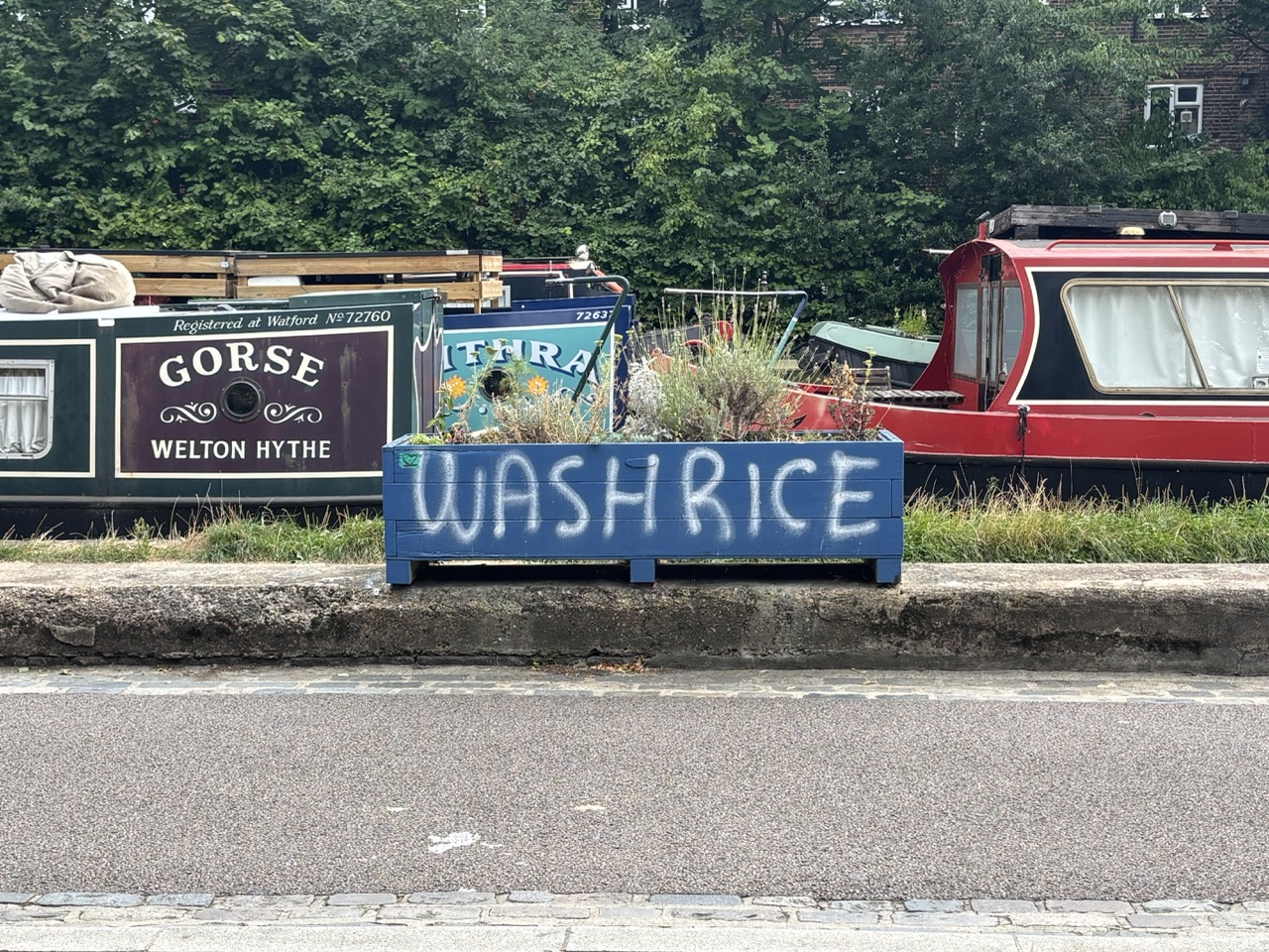 A photo of a blue planter with white spray-painted letters on it saying "Wash Rice", with boats in the background